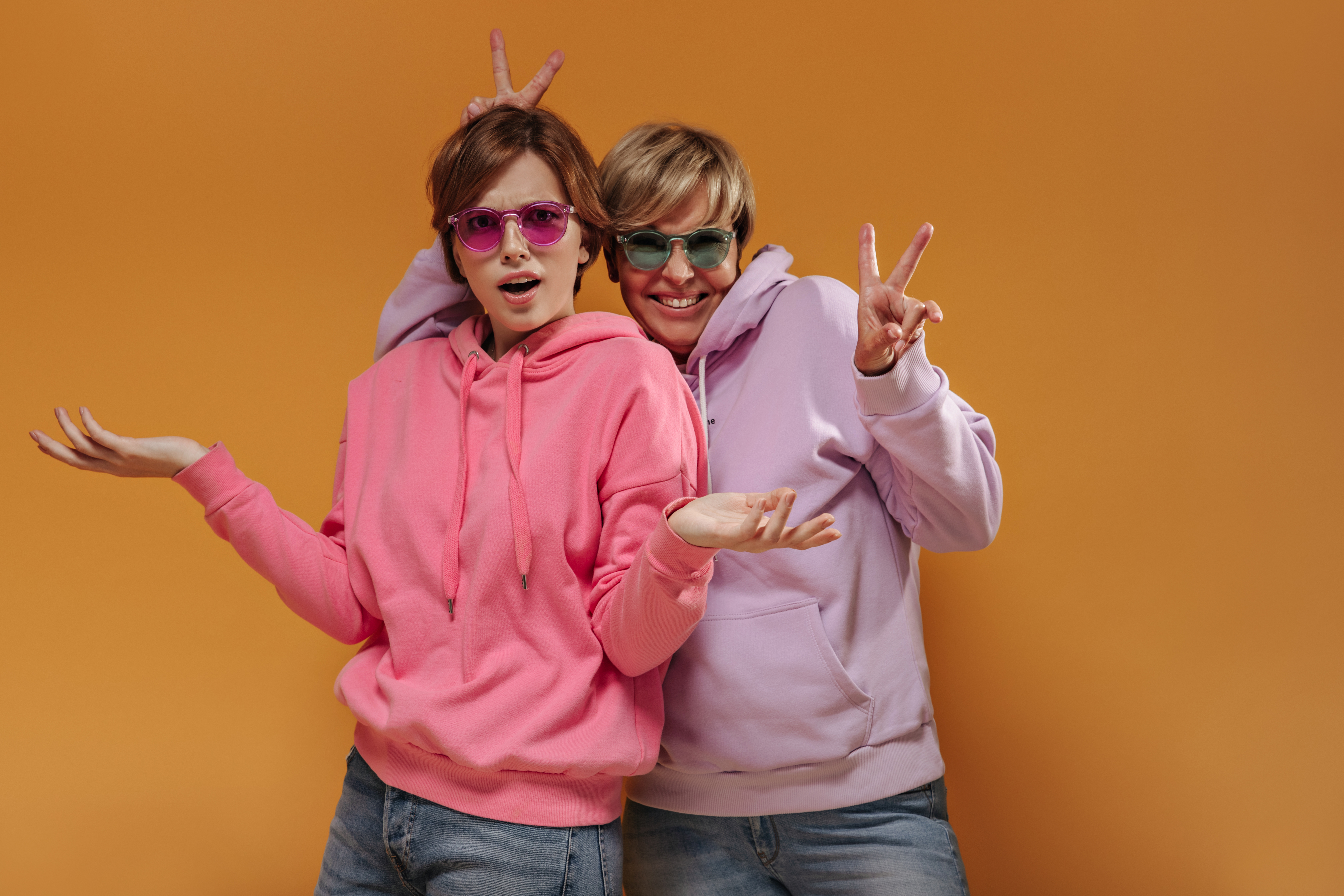 shocked-brunette-lady-sunglasses-pink-sweatshirt-looking-into-camera-posing-with-cheerful-woman-showing-peace-signs-orange-backdrop